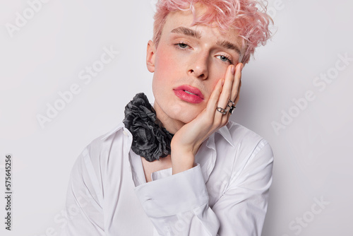 Portrait of handsome man gay with pink hair keeps hand on cheek looks attentively at camera wears white formal shirt and chain with black flower around neck isolated over white studio background