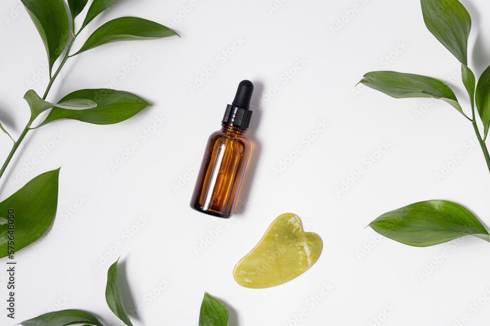 Close-up of skin serum and green quartz gouache massager on white isolated background with plant leaves. Cosmetic procedures for the face and natural skincare