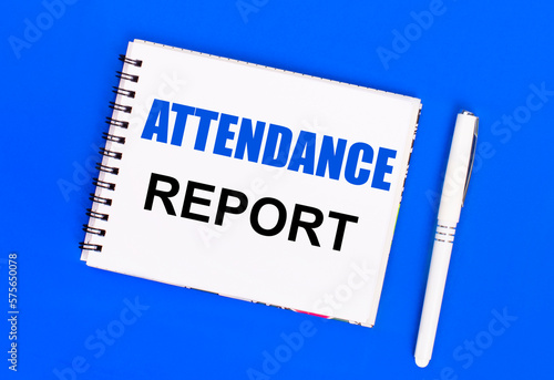 On a blue background, a white ballpoint pen and a white notepad with the text ATTENDANCE REPORT. View from above photo