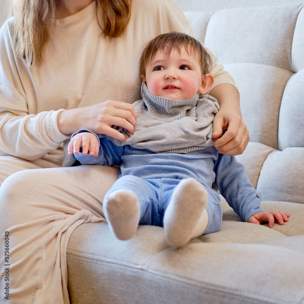 Child is dressed in warm winter clothes for a walk outside. Woman mother puts clothes on toddler baby boy for cold weather. Kid age one year five months in the home living room Child is dressed in warm winter clothes for a walk outside. Woman mother puts clothes on toddler baby boy for cold weather. Kid age one year five months in the home living room