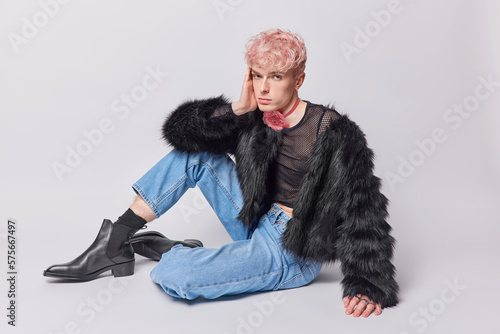 Full length horizontal shot of serious pink haired gay dressed in black fur coat jeans and leather shoes has non traditional orientation isolated over white studio background. Sexy glamour trans man