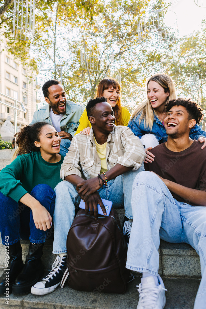 Obraz premium Vertical shot of happy multi-ethnic group of young hipster diverse student friends having fun together while hanging out sitting together outdoors. Friendship concept
