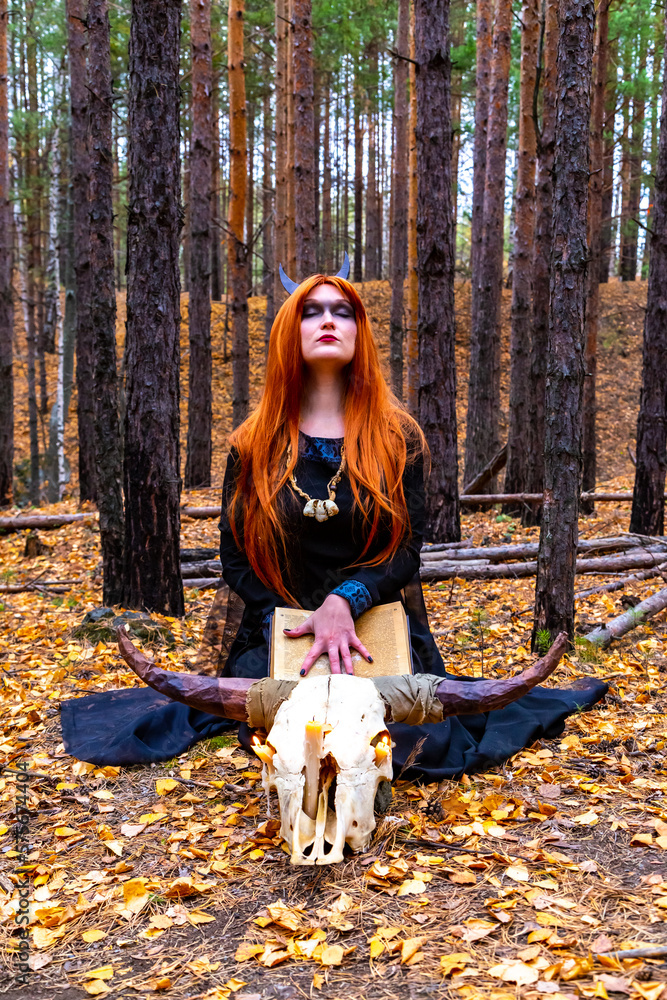 A fantastic Gothic woman, a dark witch. A red-haired evil demon girl in a black dress with a cape hood. Long hair flutters in the wind. The dark dense deep autumn forest turns the trees orange. A fantastic Gothic woman, a dark witch. A red-haired evil demon girl in a black dress with a cape hood. Long hair flutters in the wind. The dark dense deep autumn forest turns the trees orange.