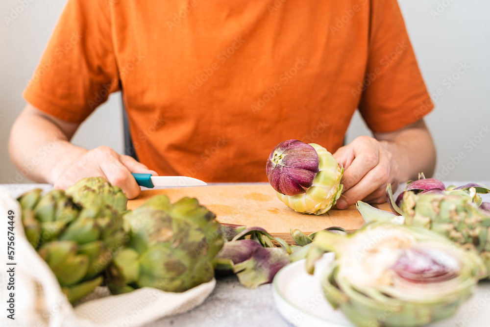 Close up Man with knife cutting artichoke on wooden cutting board on table. Cooking Healthy food and sliced vegetables artichokes. 