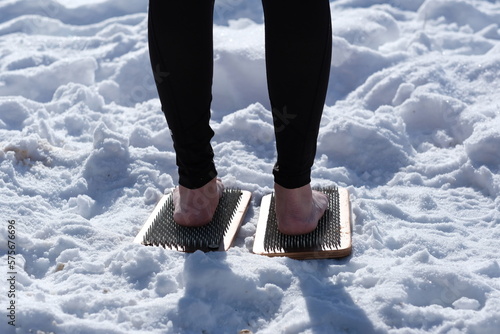 A woman practicing yoga therapy stands on a board with nails against a background of snow.