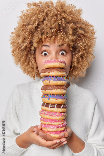Fotografija Photo of shocked curly haired woman holds stack of delicious appetizing doughnuts eats harmful food stares bugged eyes dressed in casual jumper isolated over white background