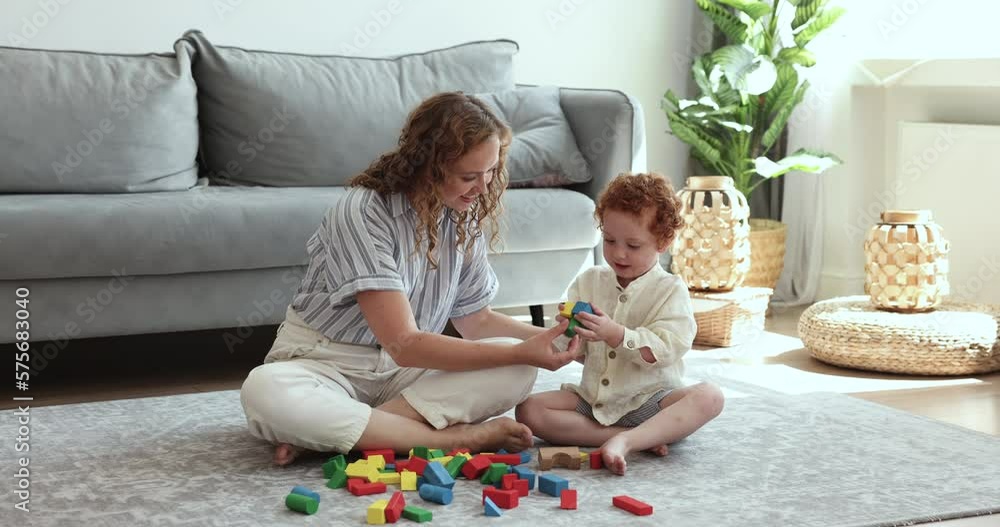 Loving young mother play with little 2s son, sit on warm floor, help teach boy build tower from developing wooden cubes, spend leisure, creative games at home. Family weekend, playtime, upbringing Loving young mother play with little 2s son, sit on warm floor, help teach boy build tower from developing wooden cubes, spend leisure, creative games at home. Family weekend, playtime, upbringing