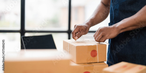Photo of young entrepreneur man packing he goods while sitting in table comfortable sitting room as background. Shipping, Shopping online, Small business entrepreneur, SME, freelance.