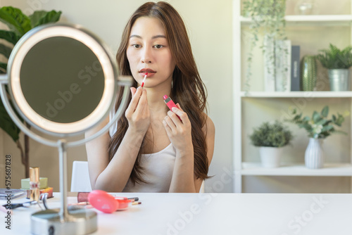 Beauty cosmetics concept, hand of asian young woman, girl make up face by applying lipstick, lips balm on her mouth, looking at the mirror at home. Female look with natural fashion style, skin care.