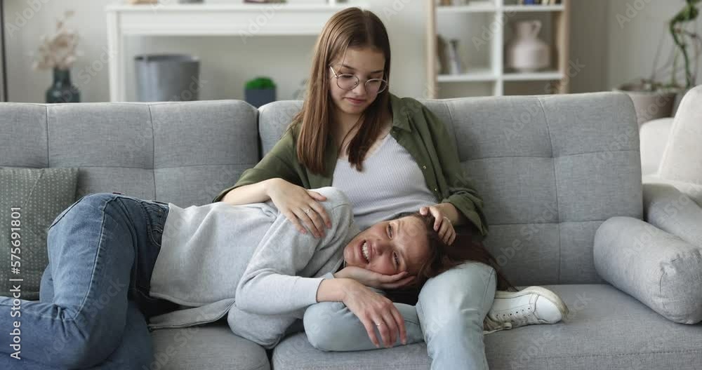 Middle-aged mother lying down on adolescent daughters knees, enjoy rest and talk. Girl strokes mom head, touch hair, takes care, feels love while relax together on couch. Joint pastime, understanding Middle-aged mother lying down on adolescent daughters knees, enjoy rest and talk. Girl strokes mom head, touch hair, takes care, feels love while relax together on couch. Joint pastime, understanding