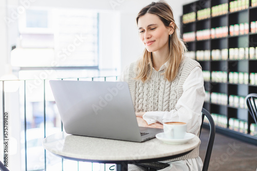 Attractive happy young girl student studying at the college library, sitting at the table with coffee, using laptop computer.Freelancer woman have video call, training coach, teacher lesson.