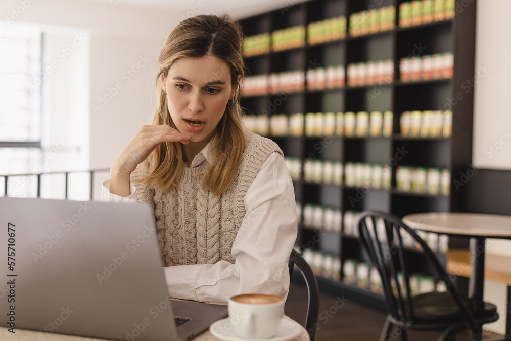 Focused young businesswoman or student looking at laptop. Serious woman working on laptop, look surprised and shock, open mouth. Girl sitting in cafe with cup of coffee. Focused young businesswoman or student looking at laptop. Serious woman working on laptop, look surprised and shock, open mouth. Girl sitting in cafe with cup of coffee.
