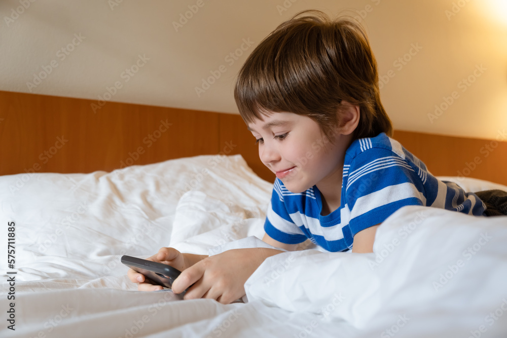 Portrait of a smiling boy playing online games on a smartphone, lying in bed at home. Child is using a mobile phone in a hotel room. Concept of gambling addiction. A kid having fun. Gadget in hands