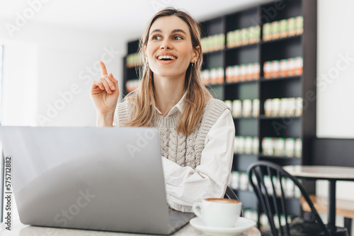 Have idea! Portrait of creative positive attractive young girl freelancer in knitted beige vest and white shirt is sitting in cafe and working on laptop with toothy smile and showing finger up. indoor