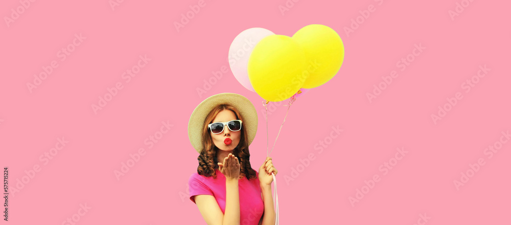Young woman with colorful balloons blowing her red lips sending sweet air kiss wearing summer straw hat on pink background