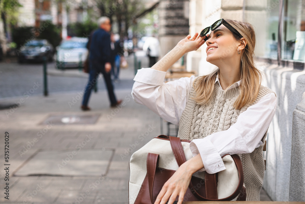 Obraz premium Young woman sitting on a street cafe waiting order, holding bag in arms and watching the city life, enjoy Europe lifestyle. Girl take off black sunglasses, wear white shirt and beige knitted vest, bag