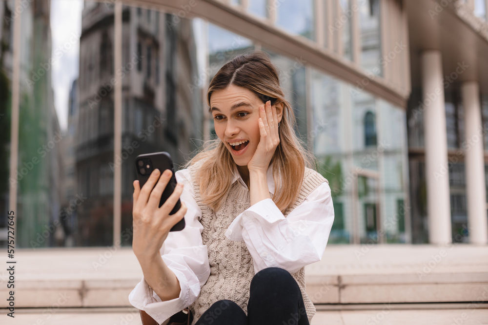 Fototapeta premium Beautiful smiling fashionable businesswoman sitting on the stairs in front of business center and make selfie, hold mobile phone, look happy. Amazed woman wear beige knit vest, white shirt and bag.