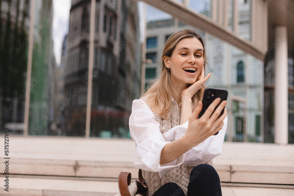 Beautiful smiling fashionable businesswoman sitting on the stairs in front of business center and make selfie, hold mobile phone, look happy. Amazed woman wear beige knit vest, white shirt and bag.