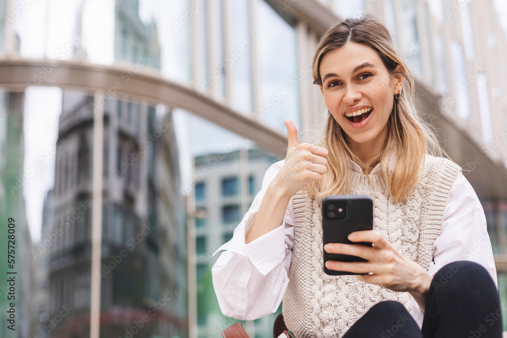 Beautiful smiling fashionable businesswoman sitting on the stairs in front of business center and show thumb up, hold mobile phone, look happy. Amazed woman wear beige knit vest, white shirt and bag. Beautiful smiling fashionable businesswoman sitting on the stairs in front of business center and show thumb up, hold mobile phone, look happy. Amazed woman wear beige knit vest, white shirt and bag.