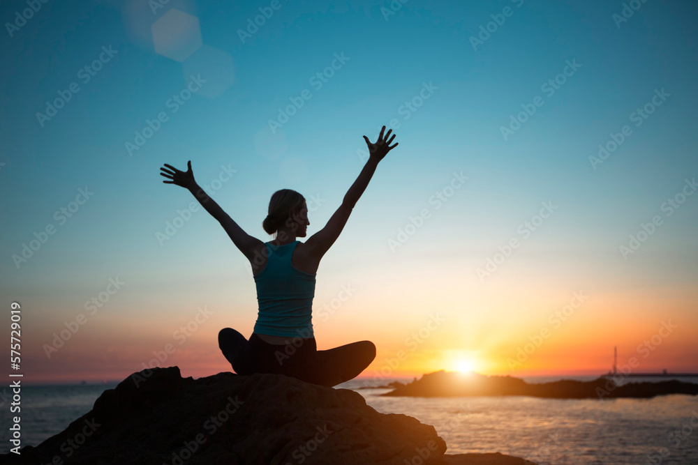 A silhouette of a woman in lotus pose doing yoga on the oceanfront during a magical sunset.