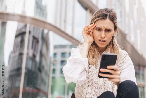 Sad being victim of cyber bullying online, anxious women checks the phone, reads a message on the stairs outside from the business center. Woman shocked and clutched her head.