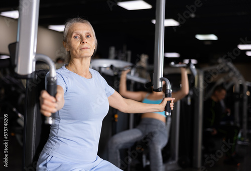 Concentrated motivated sporty elderly woman exercising chest muscles on butterfly machine during upper body strength training in gym