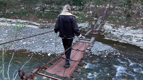 Blonde girl walks along a suspension bridge over stones and a river