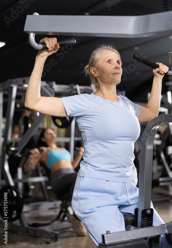 Portrait of motivated senior woman performing set of exercises for back, doing lat pull-down on lever gym machine. Concept of weight training for older adults..