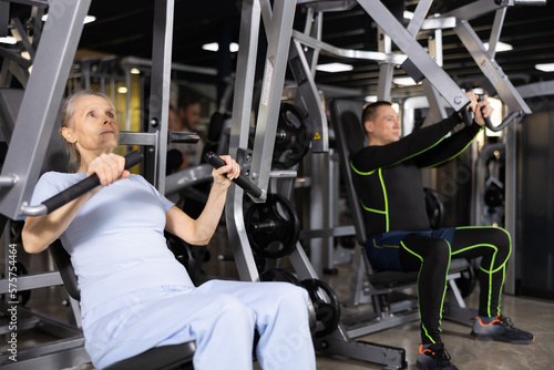 Elderly female training trains muscles of arms and press on the simulator in gym
