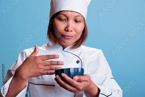 Friendly woman cook smelling healthy dish after cooking culinary meal on blue background. Positive chef wearing restaurant uniform preparing dinner. Gastronomy industry concept