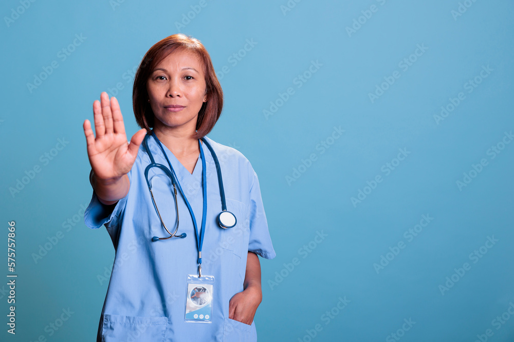 Asian medical assistant doing stop gesture with hands while working at illness expertise planning health care treatment for sick patient. Serious nurse standing in studio with blue background Asian medical assistant doing stop gesture with hands while working at illness expertise planning health care treatment for sick patient. Serious nurse standing in studio with blue background