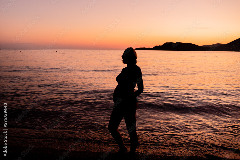 Silhouette of a pregnant woman on the seashore. Expectant mother expecting a baby. The concept of vacation, travel in pregnancy. Beautiful view of the outlines of the mountains in the orange sunset.