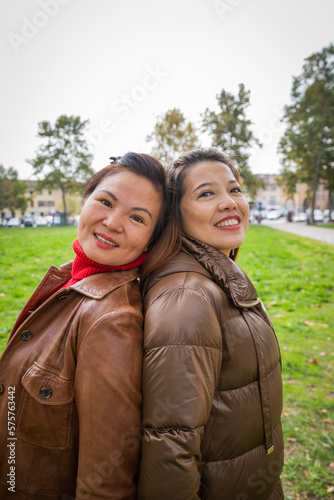 Two smiling friendly Asian women pose back to back in a public park