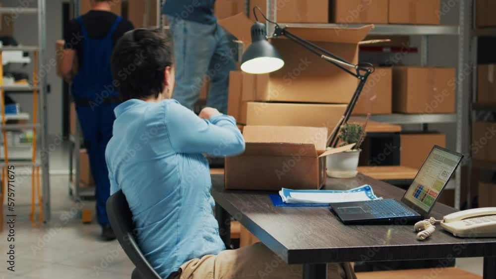 Entrepreneur using laptop to plan storage logistics, preparing merchandise order to ship to customers. Young employee putting products in packages, taking notes for stock inventory. Entrepreneur using laptop to plan storage logistics, preparing merchandise order to ship to customers. Young employee putting products in packages, taking notes for stock inventory.