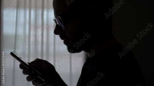 Doom scrolling. Silhouette of man scrolls page app on mobile phone. Browsing internet or social network. Compulsive scrolling concept.Side cinematic view