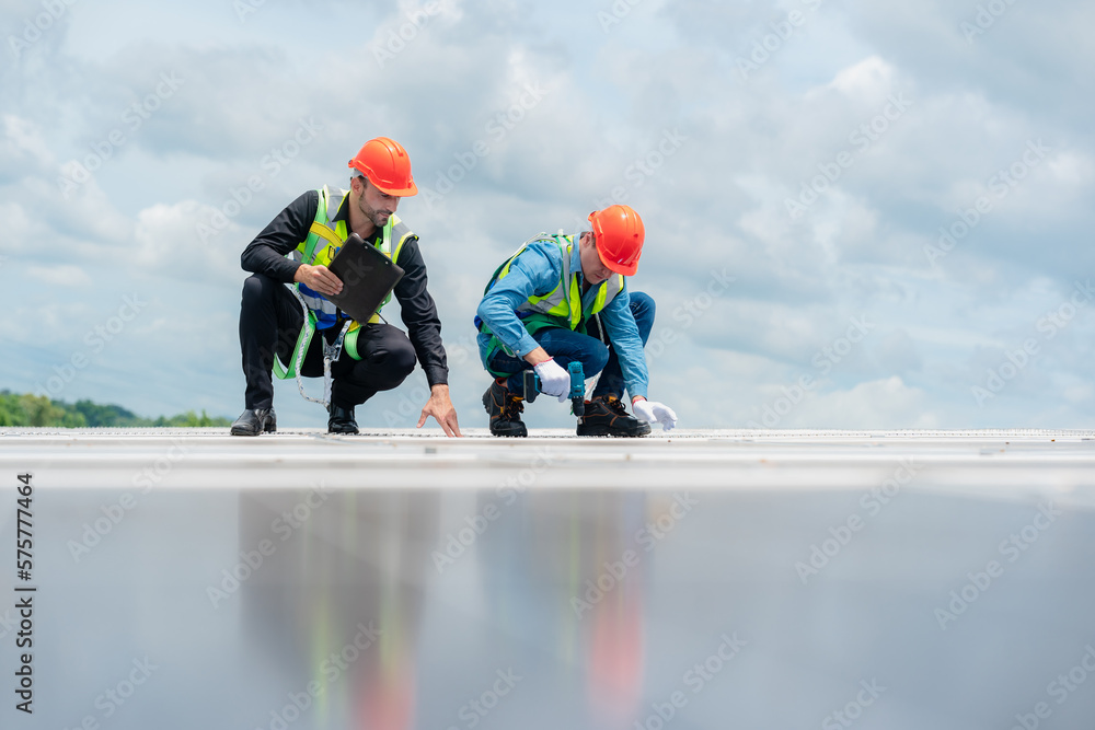 Two young engineer and inspector wearing safety vest and hard helmet inspecting solar panel by kneeling and filling holes and fixing damage using a drill machine while holding a clipboard Two young engineer and inspector wearing safety vest and hard helmet inspecting solar panel by kneeling and filling holes and fixing damage using a drill machine while holding a clipboard