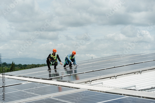 Two young engineer and inspector wearing safety vest and hard helmet inspecting solar panel by kneeling and filling holes and fixing damage using a drill machine while holding a clipboard