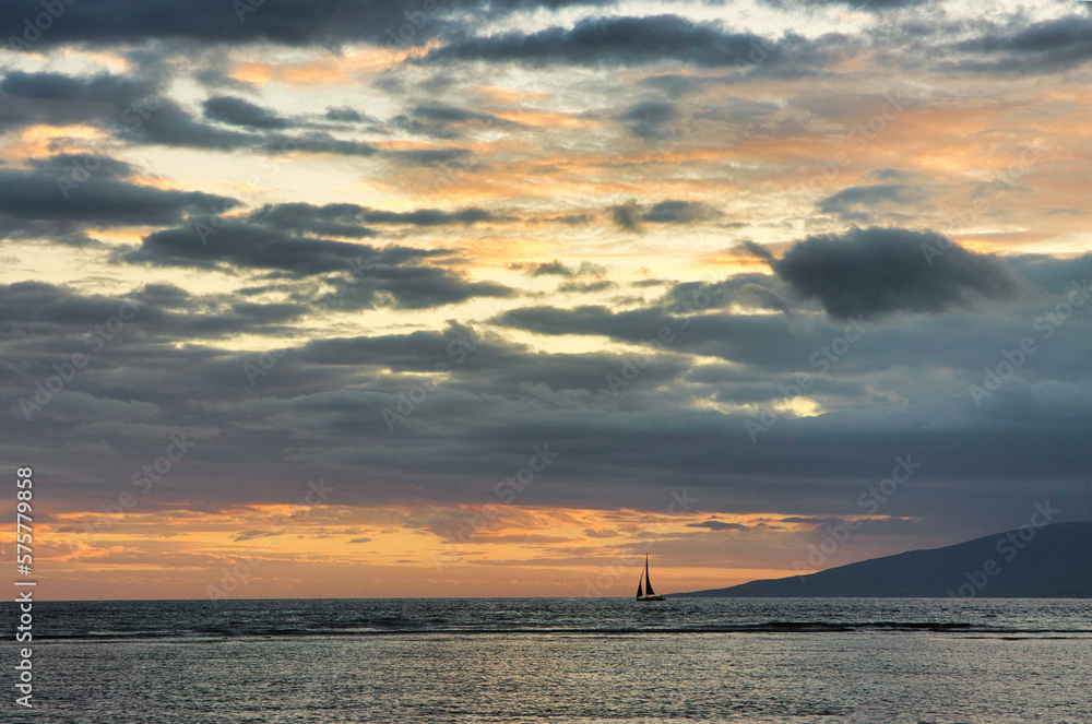 Sky ablaze during sunset on maui with a lone sailboat on the horizon.