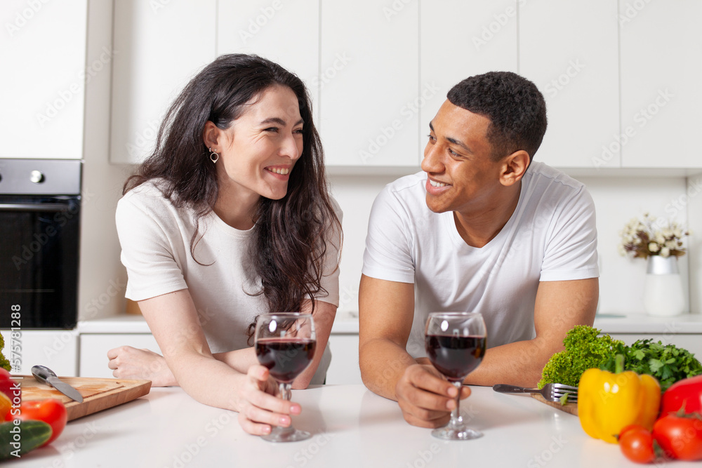 multiracial couple drinking wine in the kitchen and talking, american guy and european girl preparing salad together multiracial couple drinking wine in the kitchen and talking, american guy and european girl preparing salad together