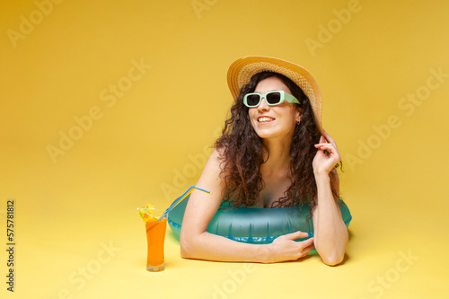 young girl in a straw hat lies on an inflatable swimming circle with an orange cocktail on a yellow background