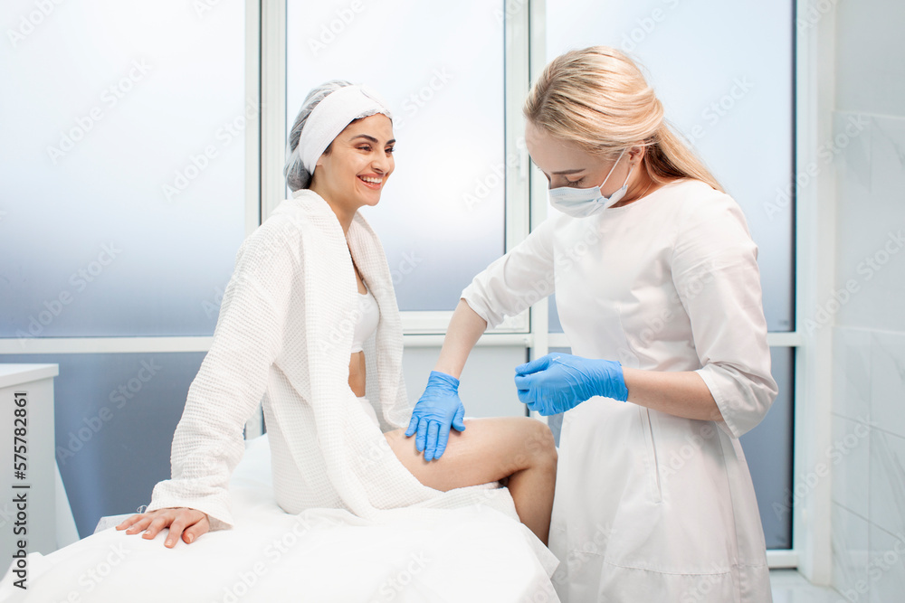 woman patient with cosmetologist doctor in cosmetology office, cosmetologist doctor applies cream on girl's leg woman patient with cosmetologist doctor in cosmetology office, cosmetologist doctor applies cream on girl's leg