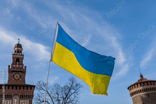 Flag with yellow and blue striped colors of Ukraine waving in the wind with a blue sky and sun. In the background the Castello Sforzesco in Milan.