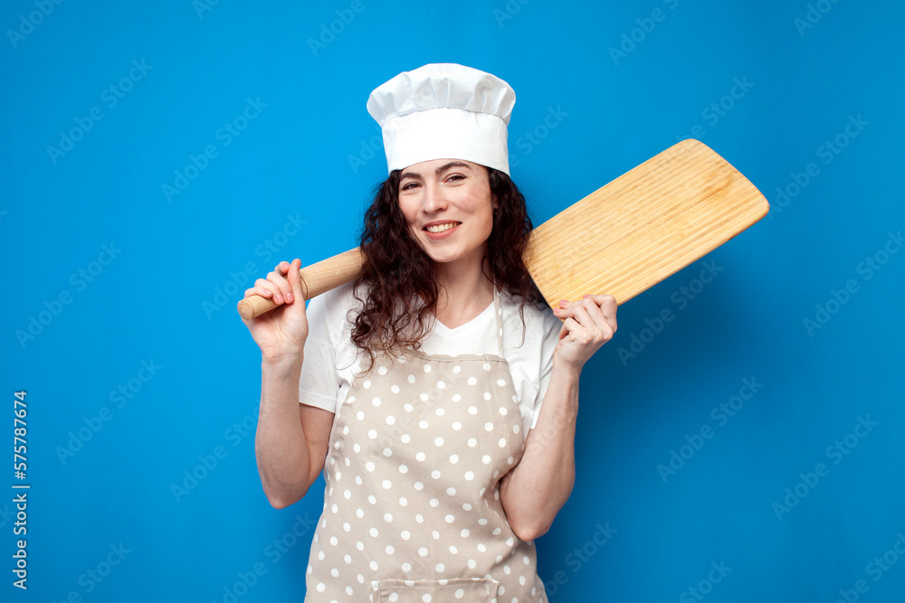 young girl chef in uniform holds kitchen item on blue background, woman housewife in an apron and hat young girl chef in uniform holds kitchen item on blue background, woman housewife in an apron and hat