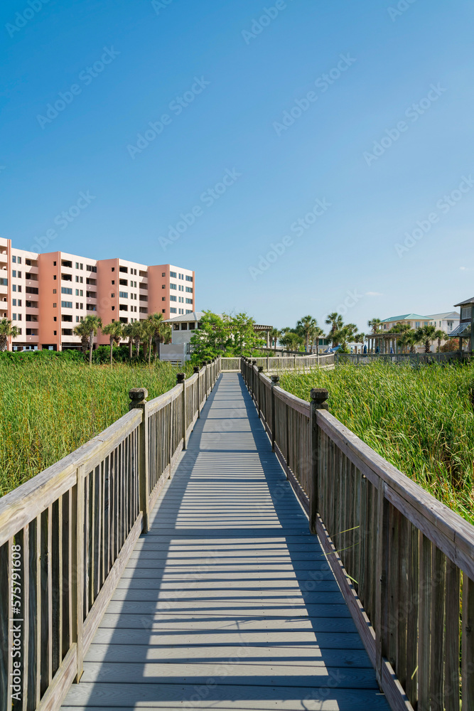 Vertical shot of a boardwalk with wooden handrails over the grasses in a neighborhood at Destin, FL. Pathway against the views of an apartment on the left and single-family homes on the right.