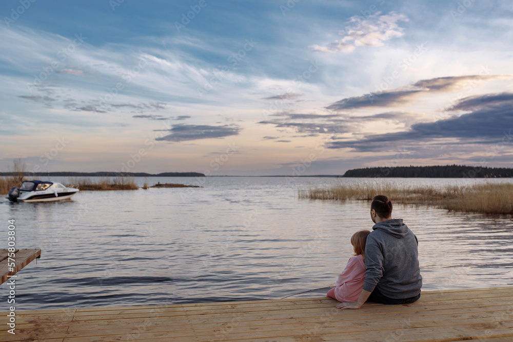 Father and daughter on the shore of the lake watching the sunset sitting on a wooden pier. Lifestyle, leisure, happiness, joy concept. Leisure.