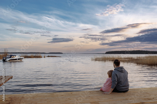 Father and daughter on the shore of the lake watching the sunset sitting on a wooden pier. Lifestyle, leisure, happiness, joy concept. Leisure.