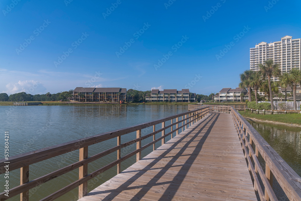 Perspective view of footbridge over Stewart Lake heading to hotels and apartments in Destin, Florida. Lakefront apartments and hotels with low-rise front and high-rise back structures against sky.