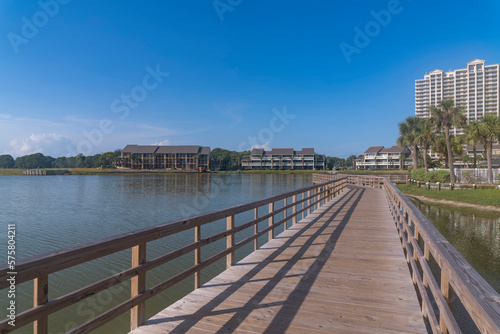 Perspective view of footbridge over Stewart Lake heading to hotels and apartments in Destin, Florida. Lakefront apartments and hotels with low-rise front and high-rise back structures against sky.