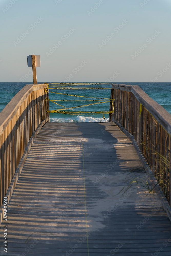 Vertical shot view of a wooden boardwalk with yellow caution tape in Destin, Florida. Walkway with wooden railings and sign post on the left and a view of blue ocean and sky background.