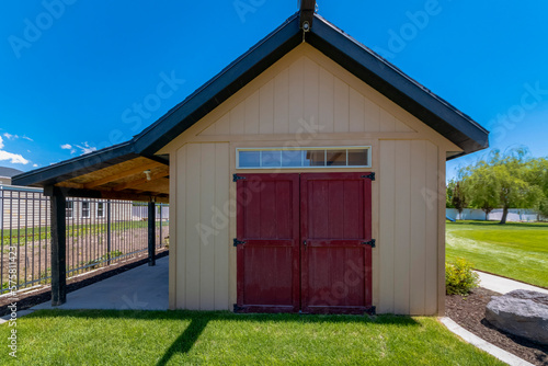 Tool shed with wood beige wall and side-hinged red double door. Shed with extended roof on the left side near the fence railings and a lawn at the front and on the right side.
